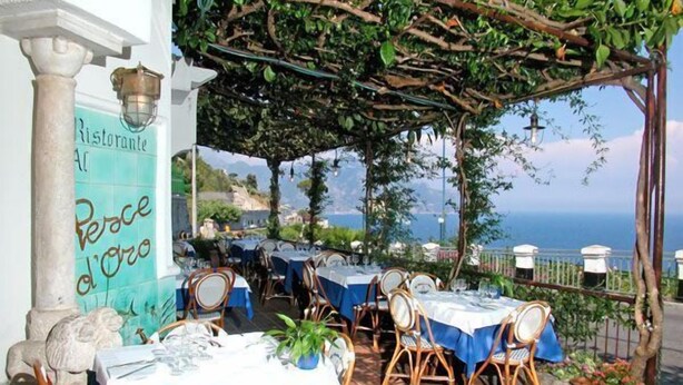 Un área de comedor al aire libre con mesas preparadas para dos, cubierta por una pérgola con plantas trepadoras, con vistas a un paisaje costero.