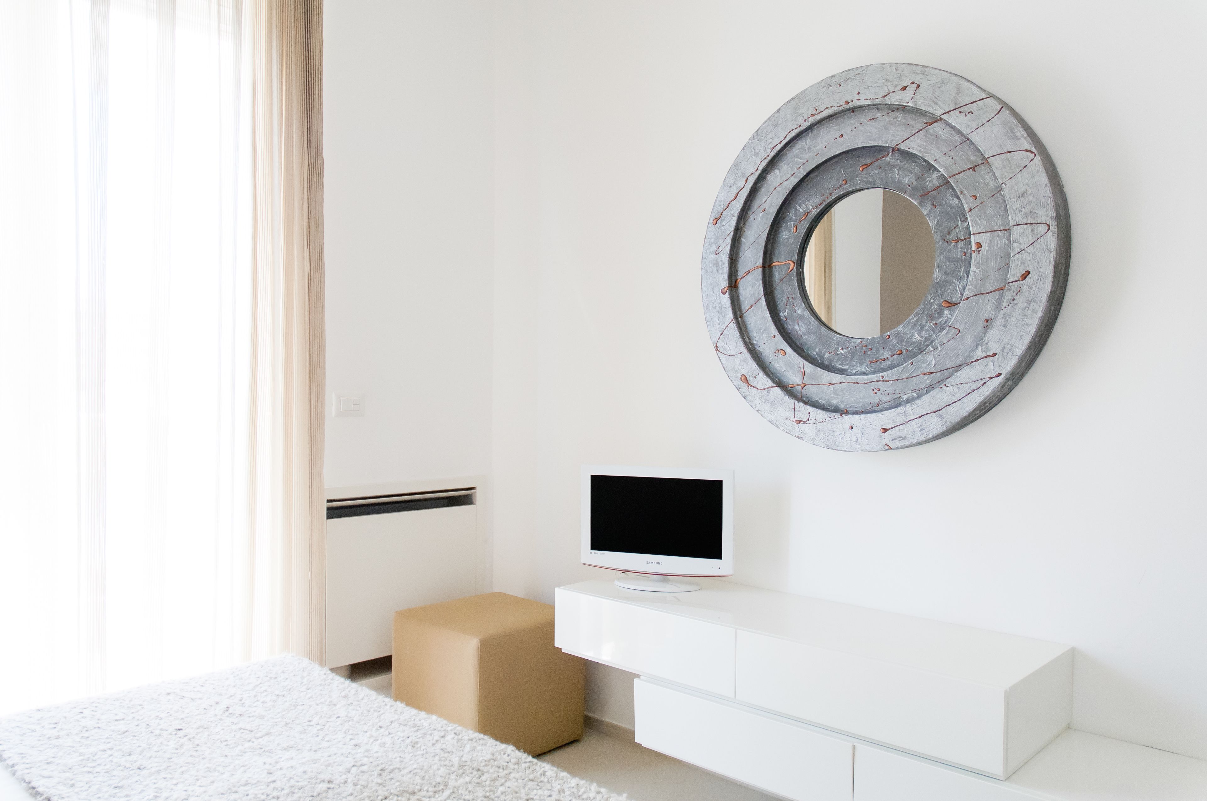 Een moderne slaapkamer met een witte dressoir, een televisie erboven en een grote ronde spiegel aan de muur.