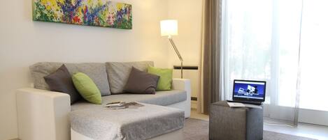 A modern living room with a grey sofa, a small ottoman, and a laptop on a side table.