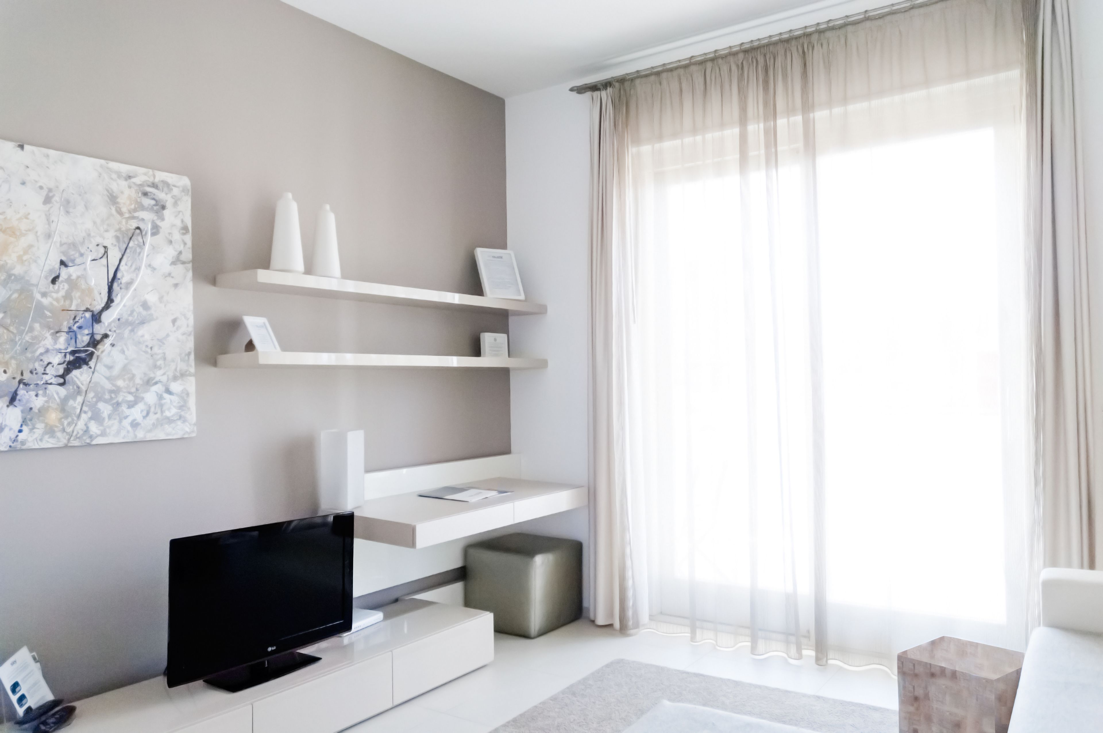Ein modernes Wohnzimmer mit Flachbildfernseher, weißen Regalen, einem grauen Hocker und einem großen Fenster mit leichtem Vorhang.