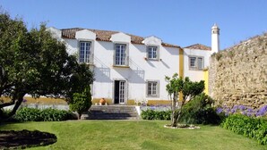 Un bâtiment à deux étages avec une façade blanche et un mur d’accent jaune, un mur de pierre sur la droite, et un petit jardin avec un arbre et des fleurs violettes.