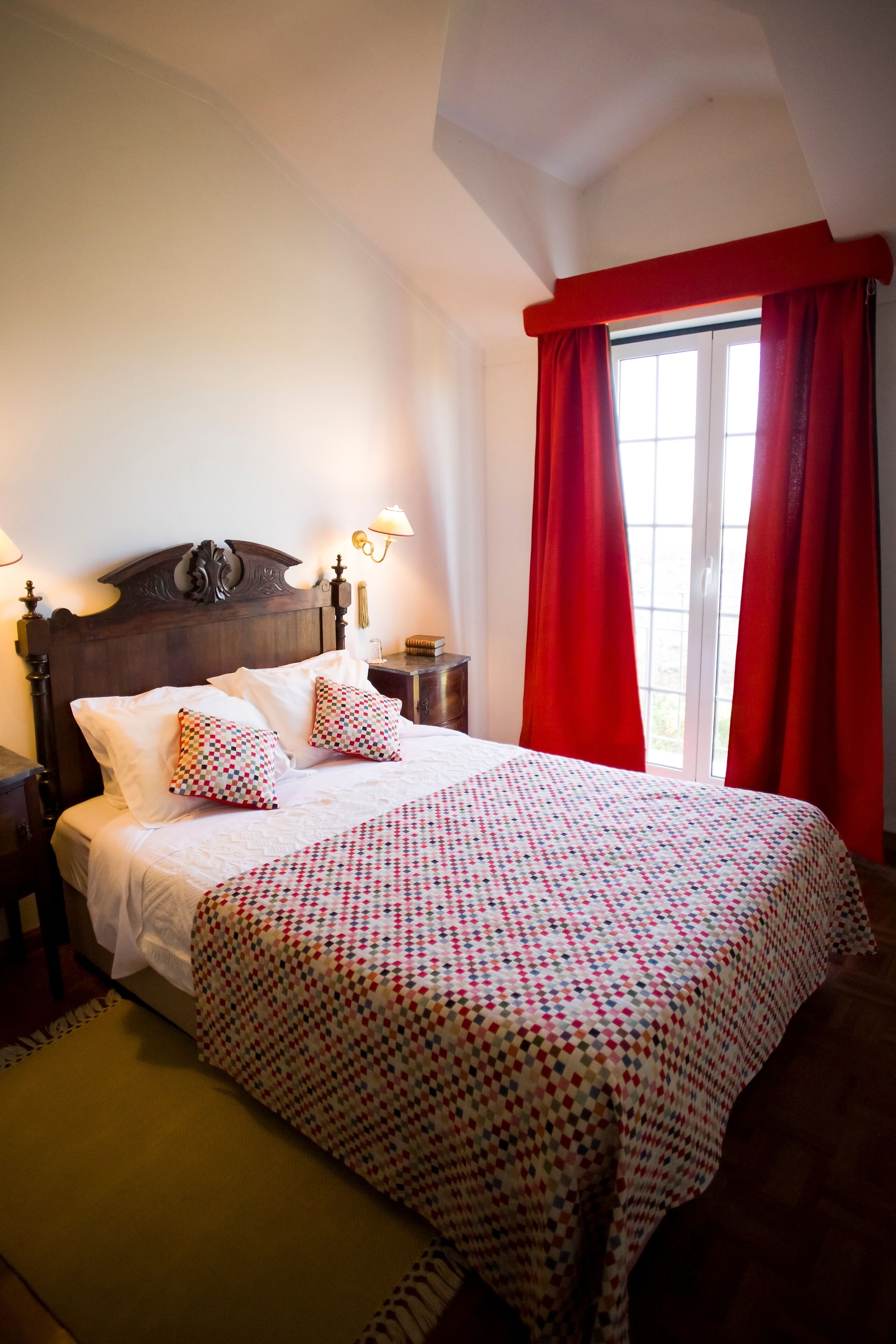 A bedroom with a wooden headboard, a bed with a checkered bedspread, two pillows, a nightstand with a lamp, and a window with red curtains.
