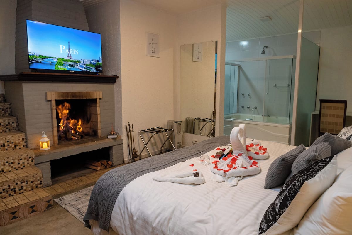 Ein Hotelzimmer mit Kamin, einem Fernseher, der eine Pariser Szene zeigt, einer Dusche sowie einem Bett mit Rosenblättern und Handtüchern.
