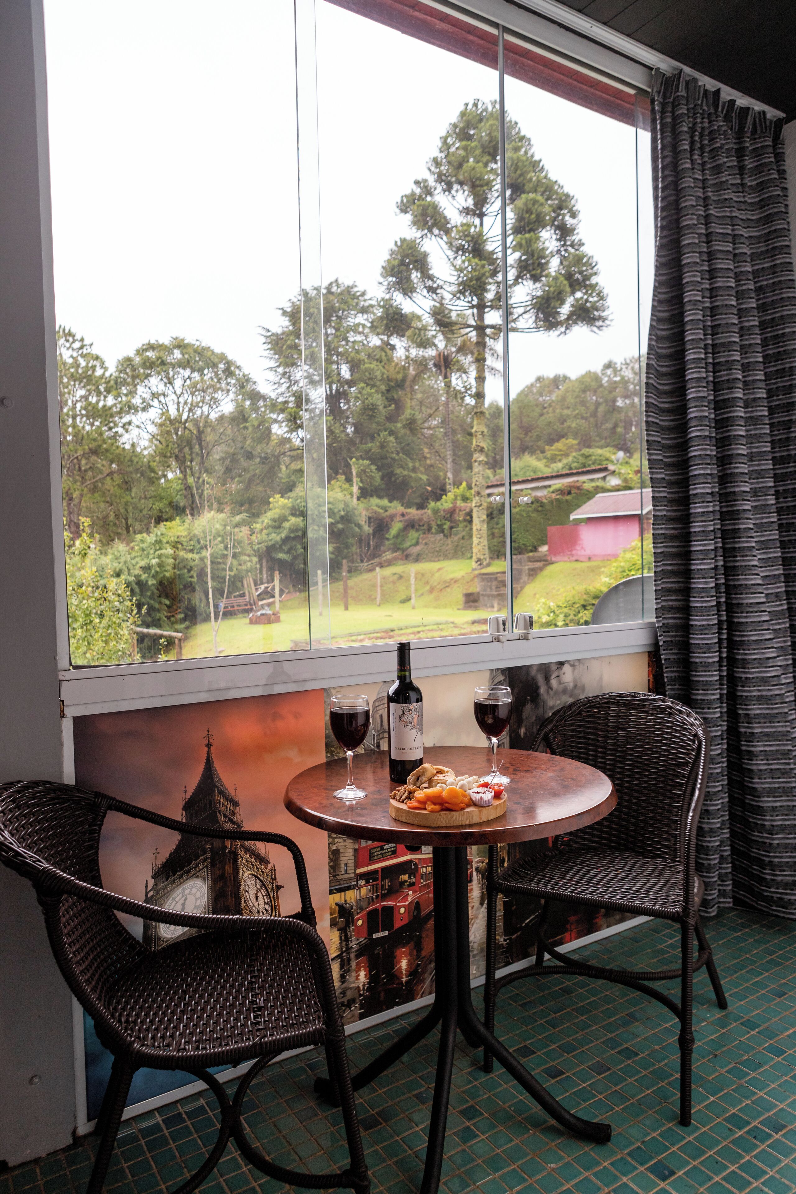 A room with a table and two wicker chairs, a window with a view of greenery and a red building, and a bottle of wine on the table.