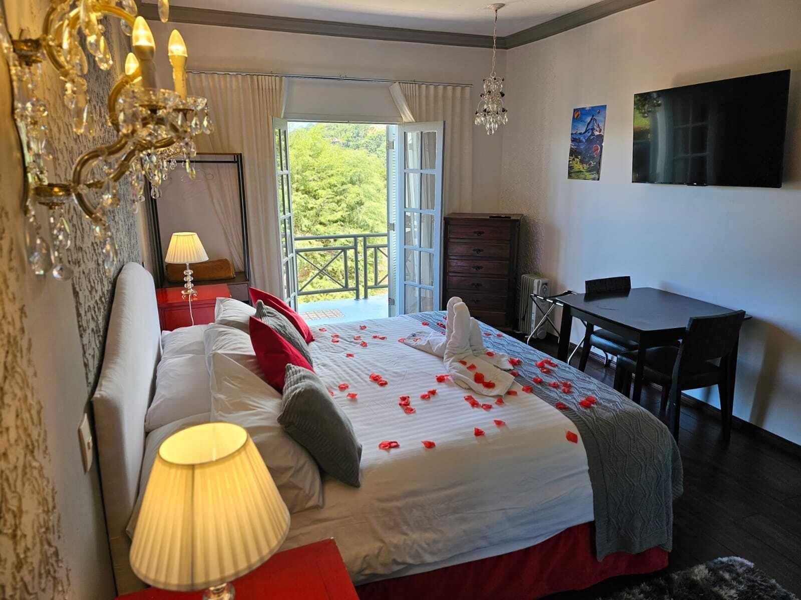 A hotel room with a bed, a desk, a TV, a chandelier, and a balcony with a view of greenery.