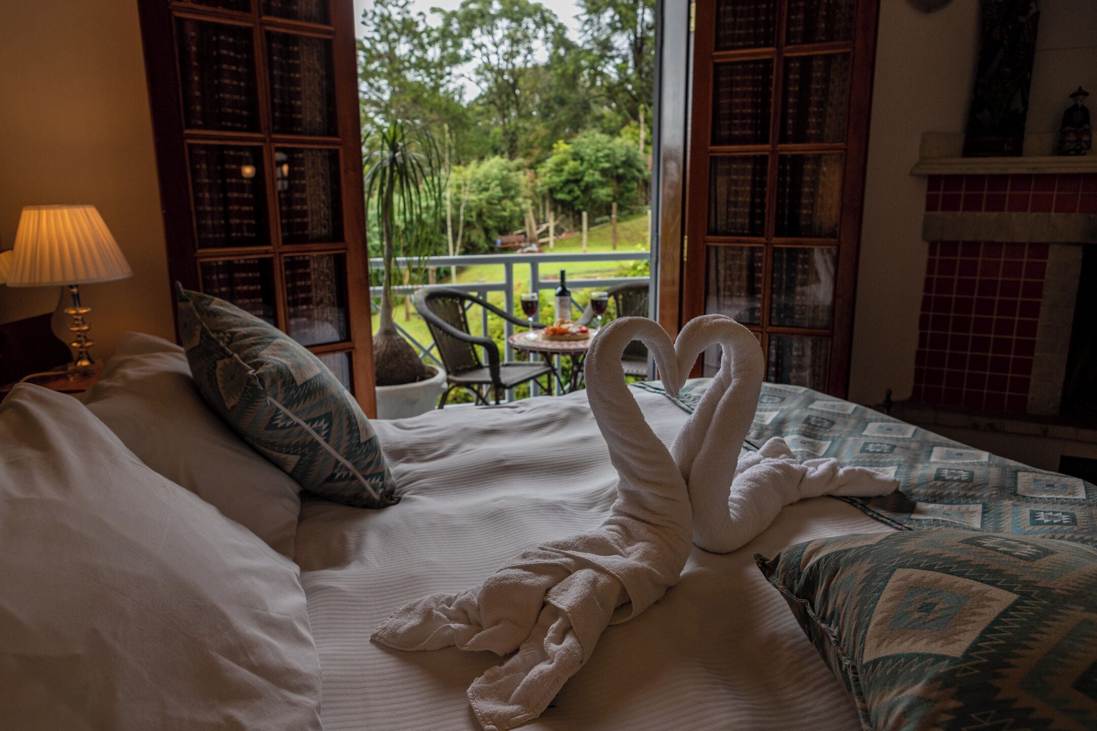 A bed with two white towels arranged in a heart shape, a lamp, and a view of a garden through a window.