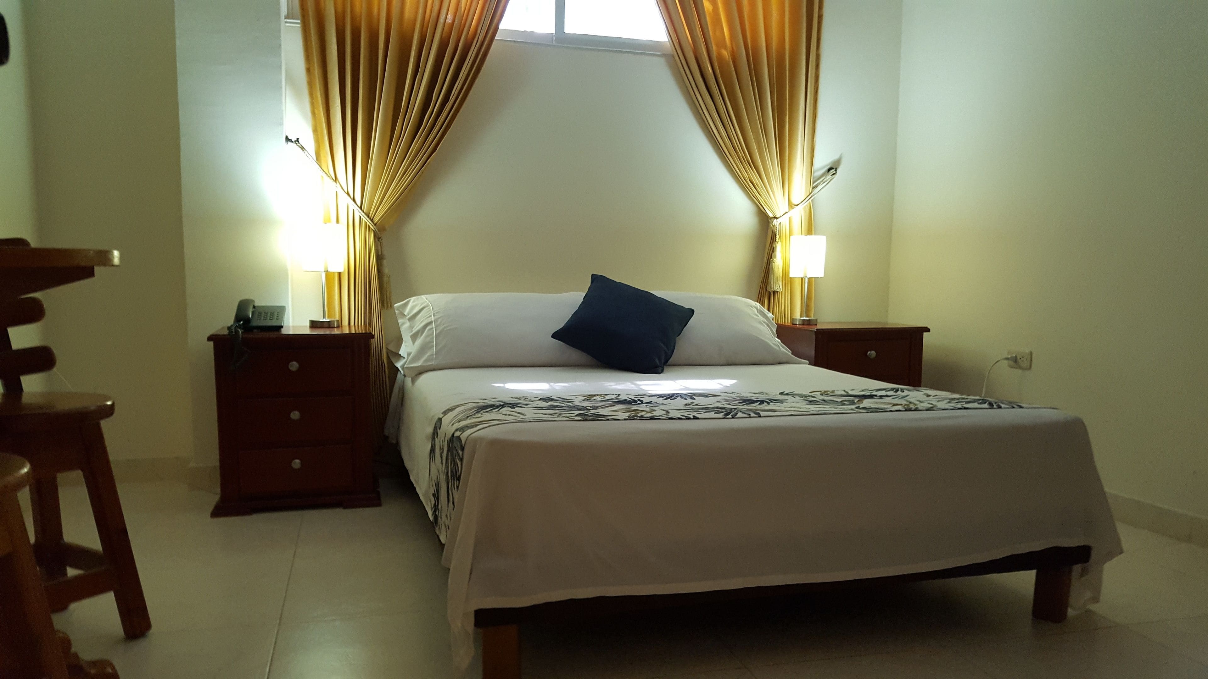 Un dormitorio con una cama, dos mesitas de noche, una butaca y cortinas.