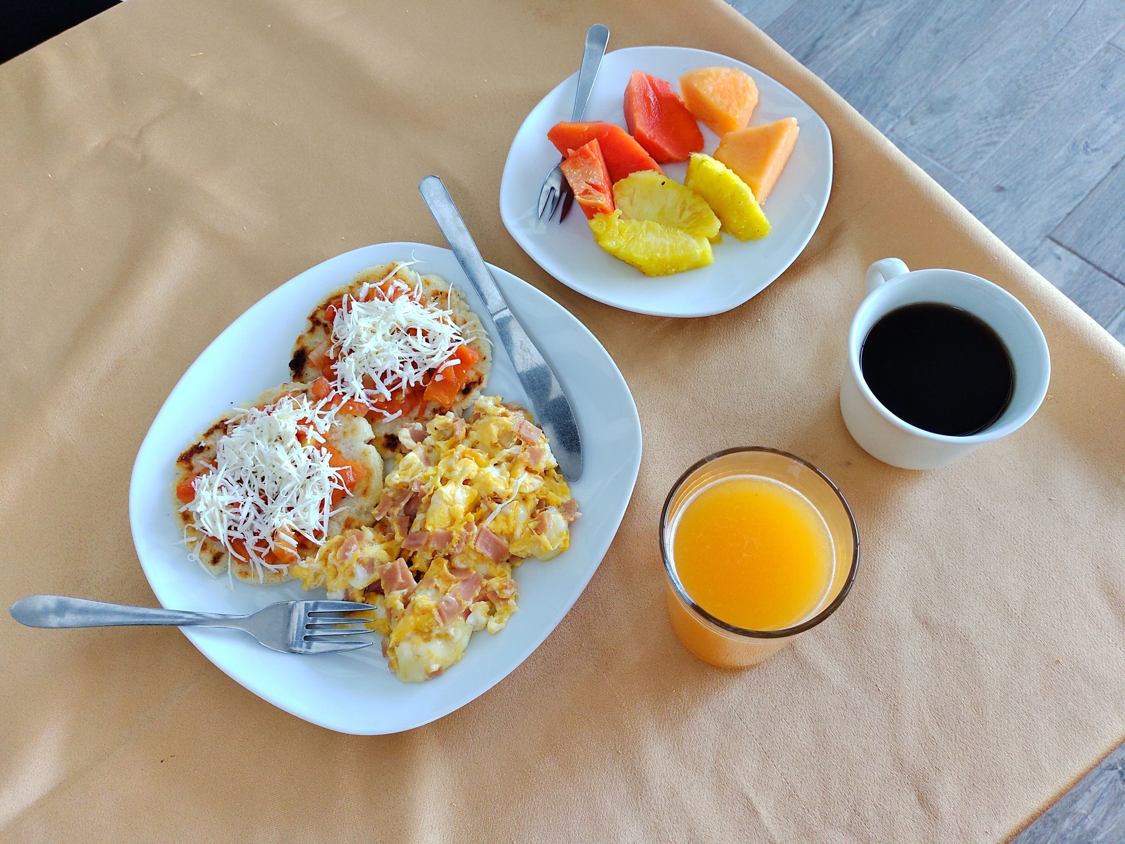 Una mesa de desayuno con huevos revueltos, queso y jamón, un plato de fruta, una taza de café y un vaso de jugo de naranja.