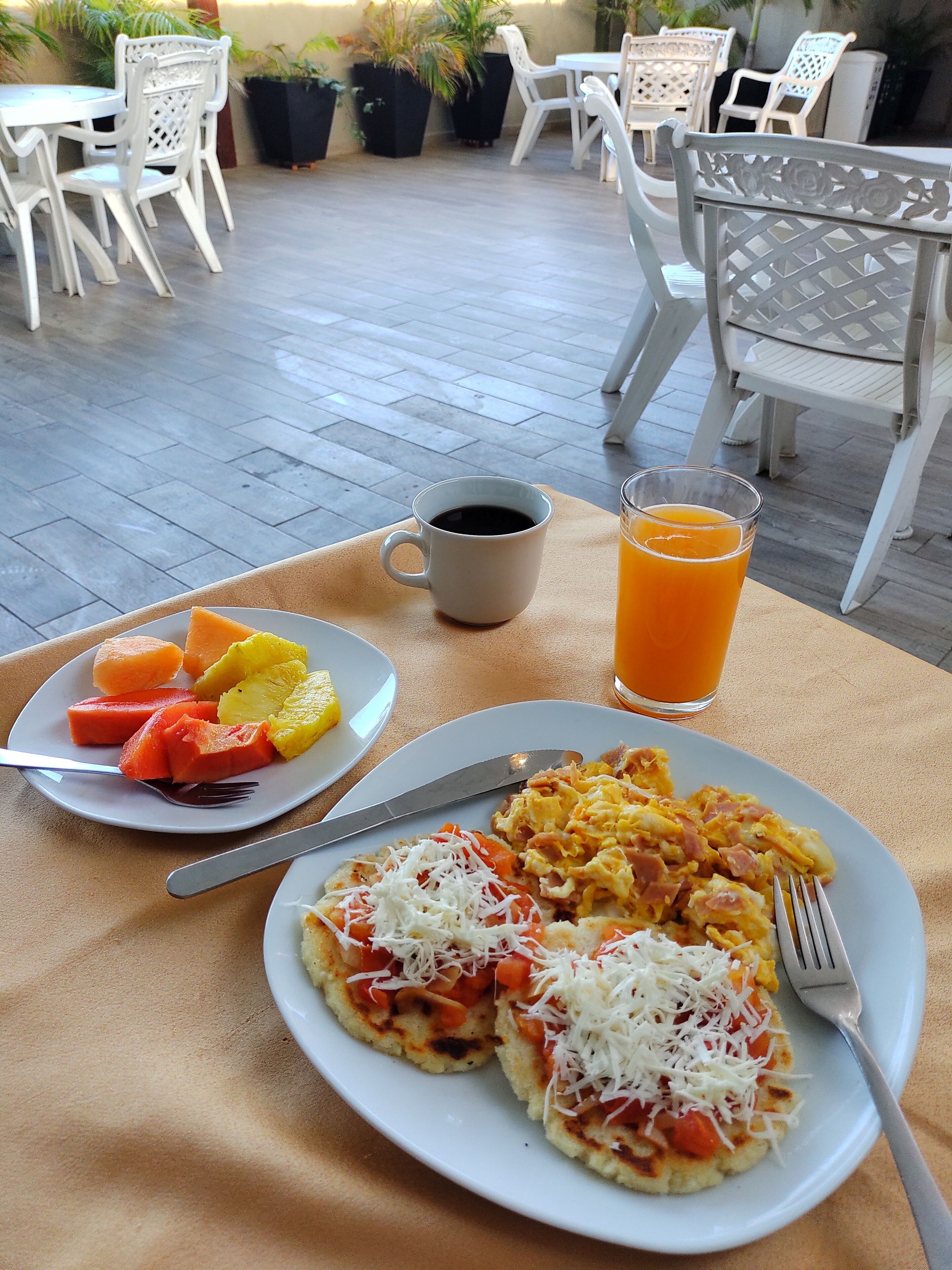 Un desayuno con platos de comida, una taza de café y un vaso de jugo de naranja sobre una mesa al aire libre.
