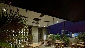 Zona pranzo all'aperto con tavoli e sedie di legno, circondata da piante in vaso e vista sulla città di notte.