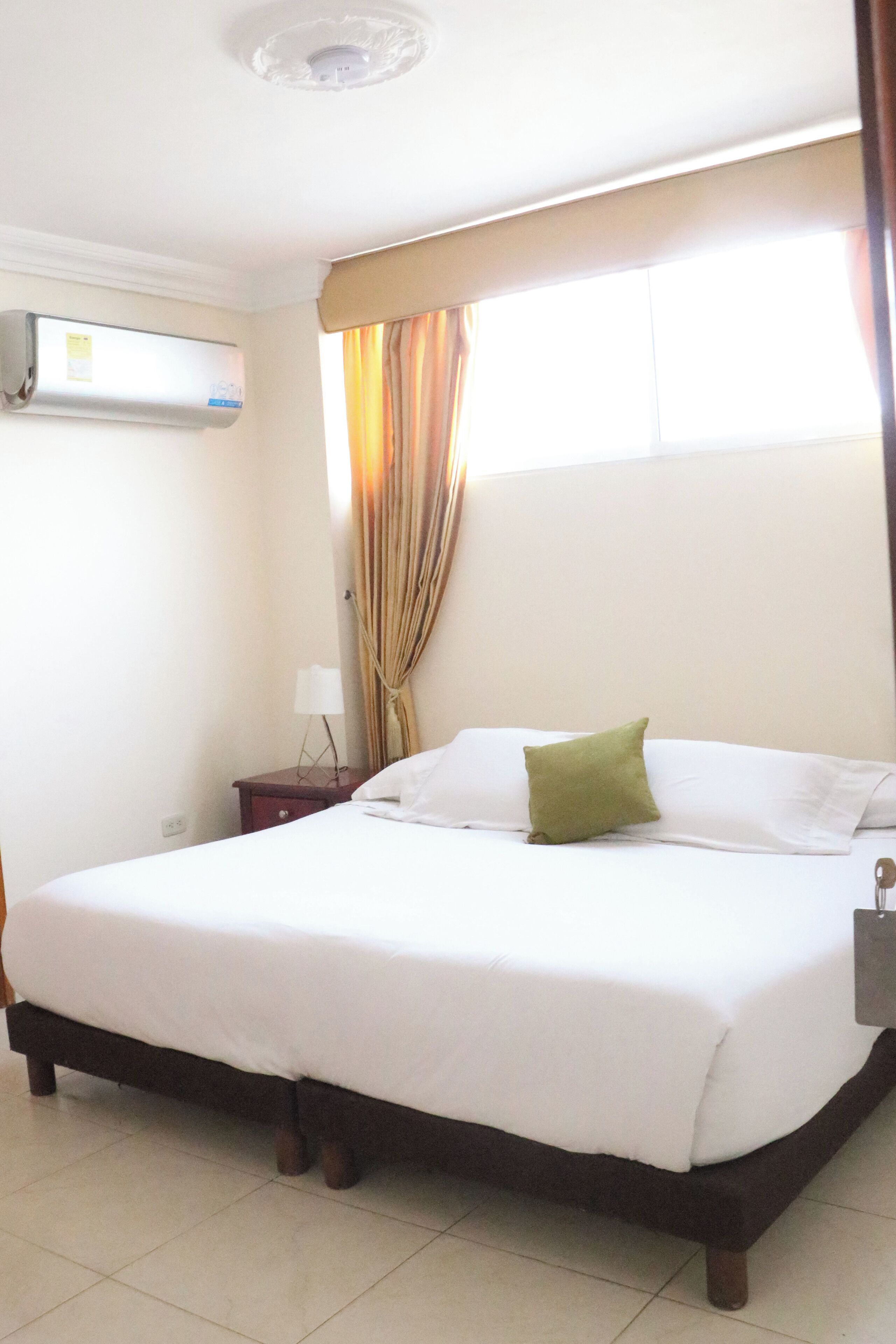 Una habitación de hotel con cama, unidad de aire acondicionado, mesita de noche, lámpara y ventana con cortinas.