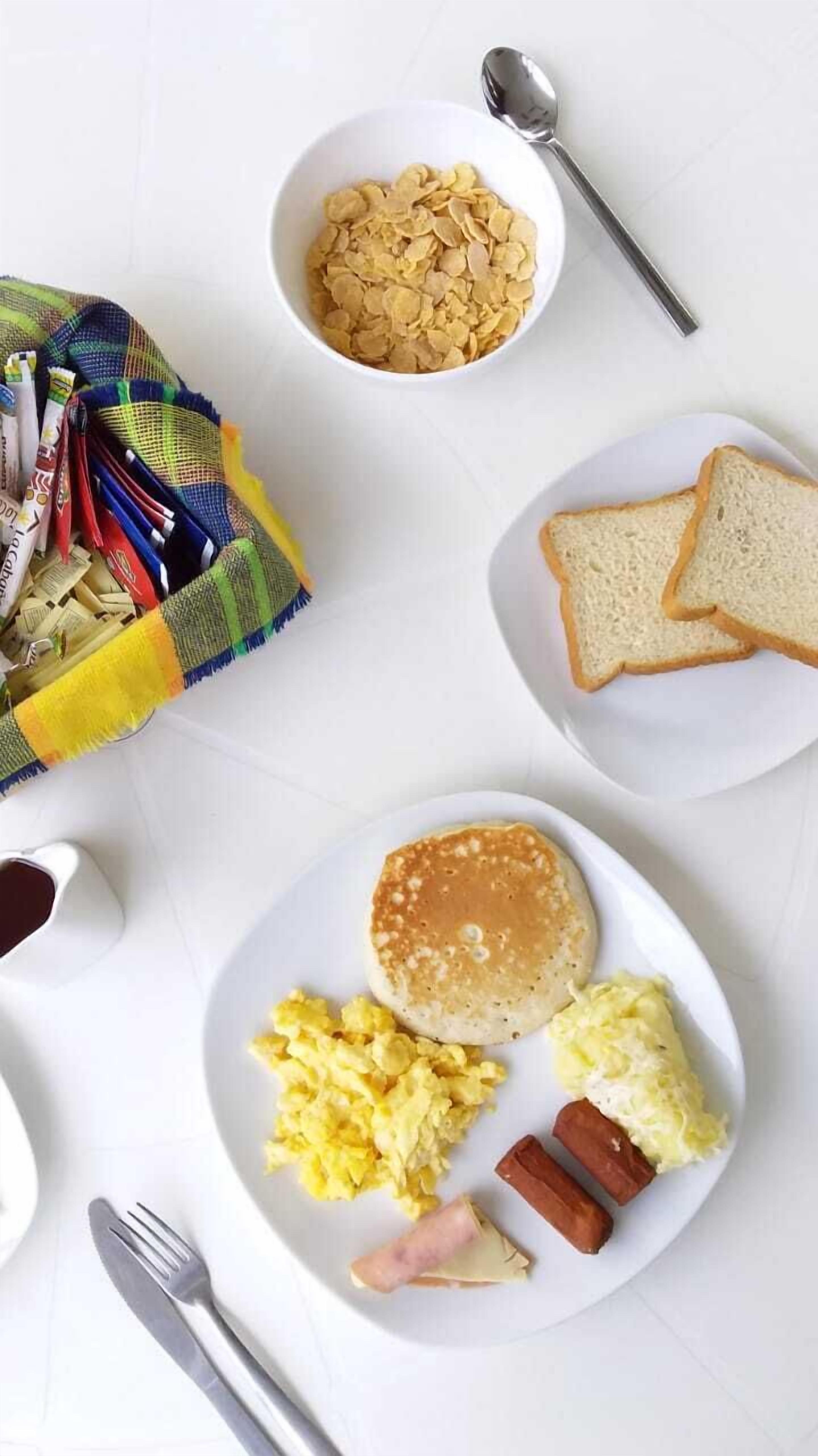 A breakfast plate with pancakes, scrambled eggs, sausages, and toast, along with a bowl of cereal and a spoon.
