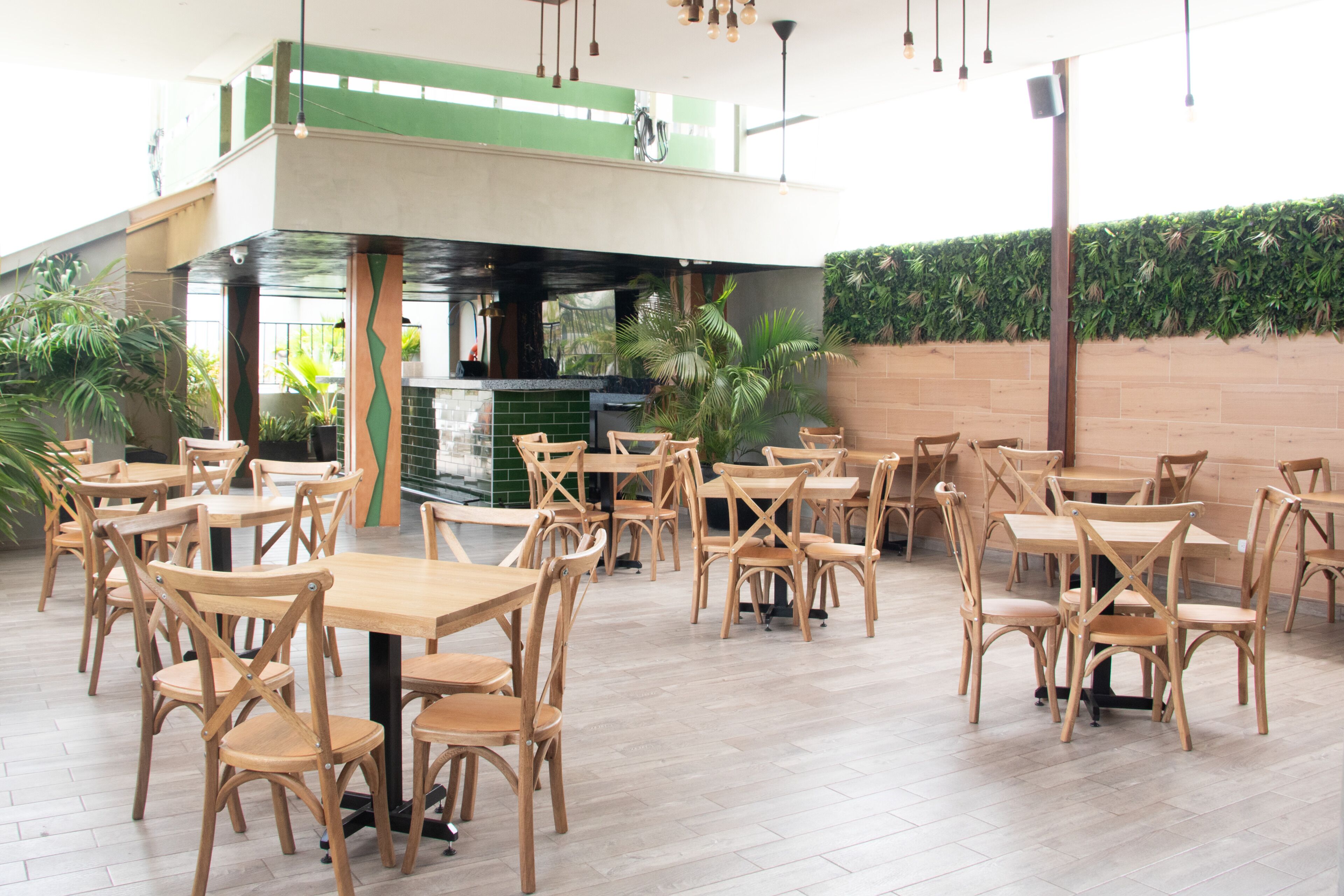 A spacious indoor dining area with wooden tables and chairs, a kitchen in the background, and a green wall with plants.
