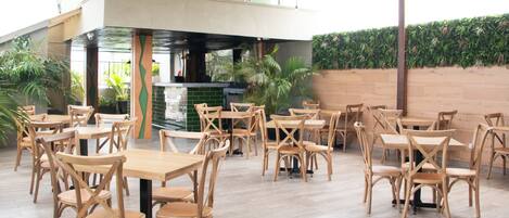 A spacious indoor dining area with wooden tables and chairs, a kitchen in the background, and a green wall with plants.