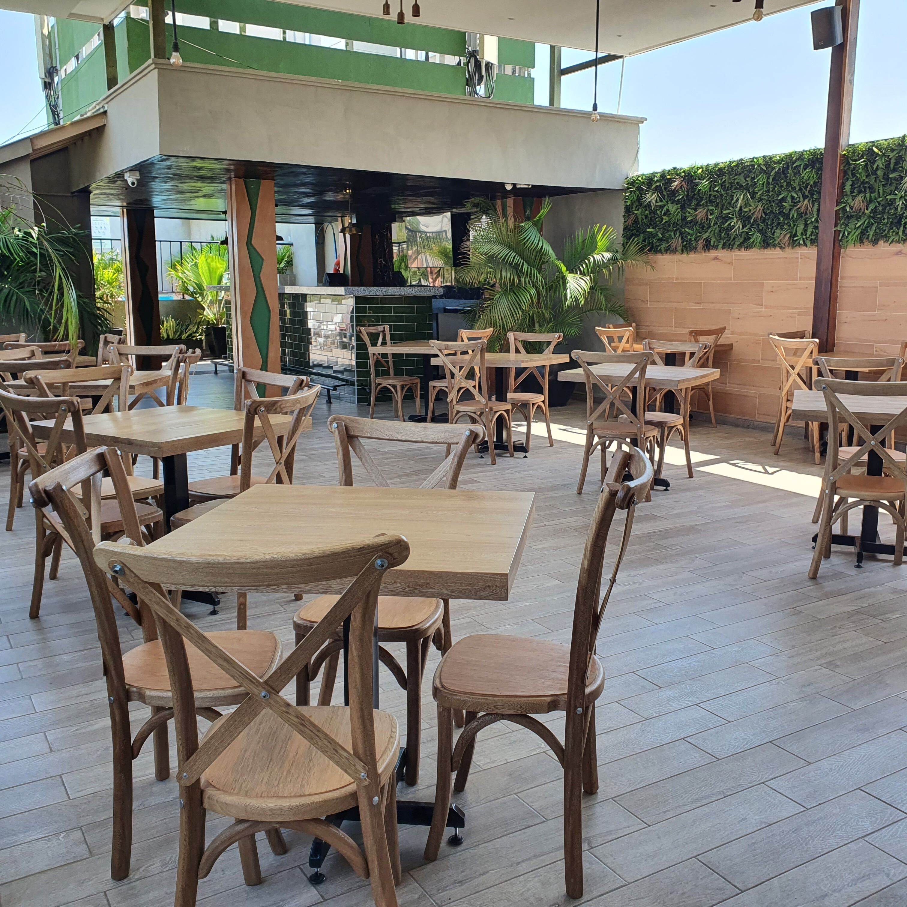 An outdoor cafe area with wooden tables and chairs, a green wall, and a tiled floor.