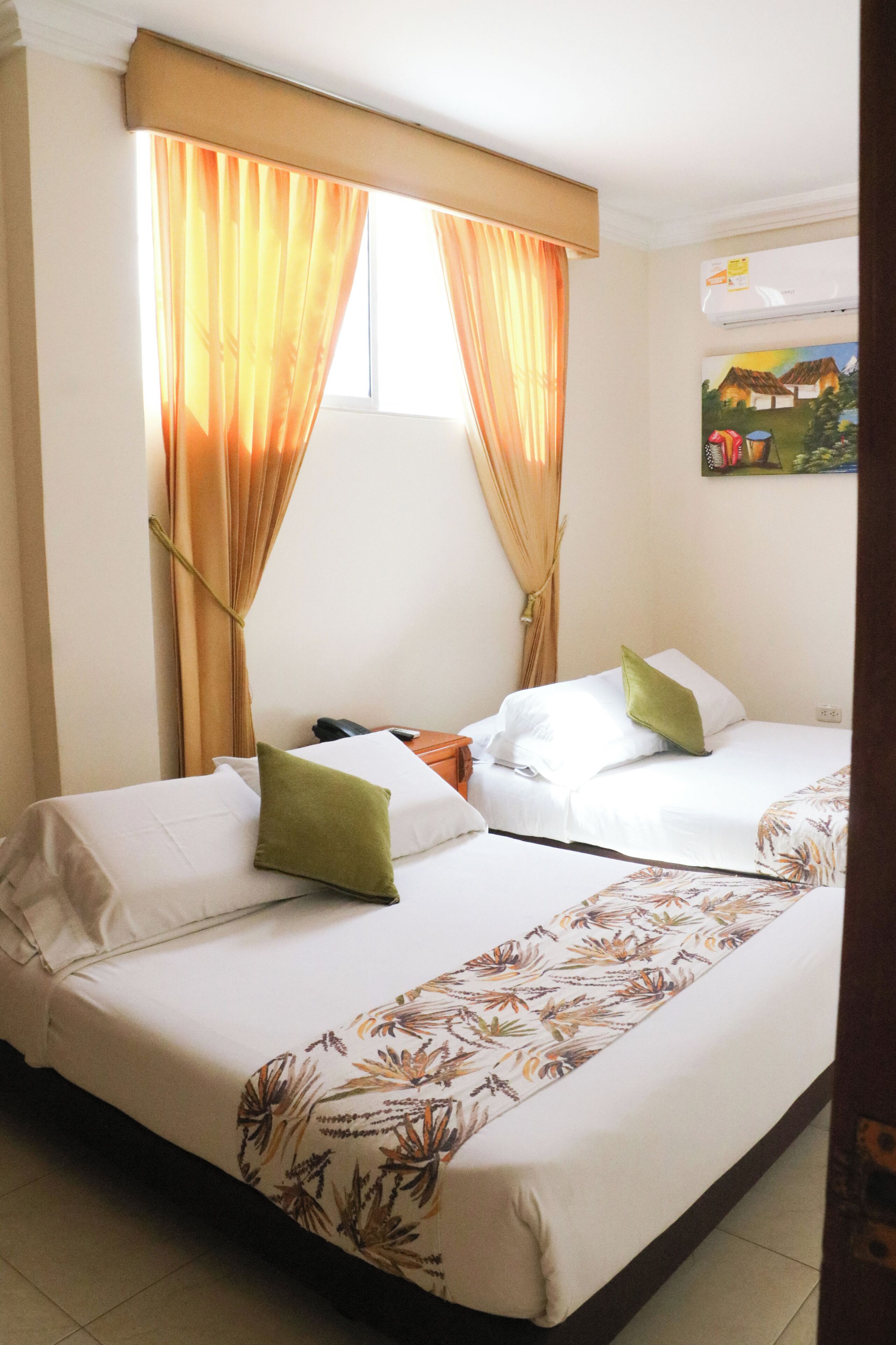 A hotel room with two beds, each with white linens and green pillows. There is a window with orange curtains, and a painting of a village on the wall.