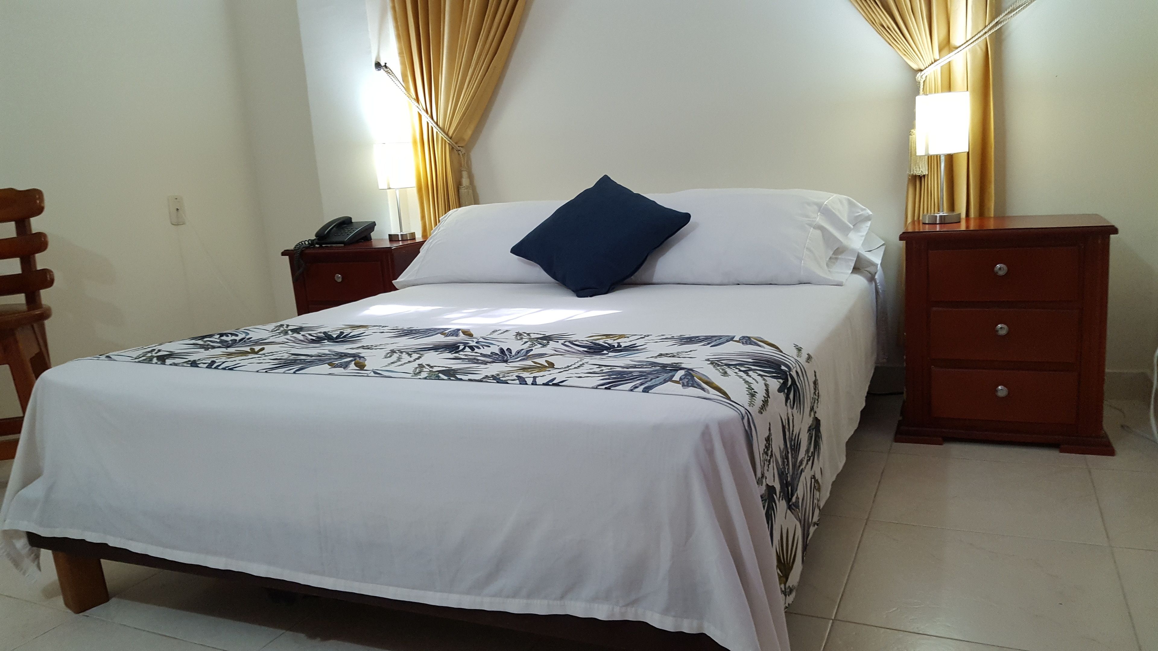 A neatly made bed with a floral bedspread, a bedside table with a lamp, and a wooden nightstand with a phone.