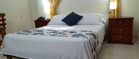 A neatly made bed with a floral bedspread, a bedside table with a lamp, and a wooden nightstand with a phone.