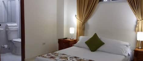Una habitación de hotel con cama, mesita de noche, lámpara de noche, ventana con cortinas y vista al baño.