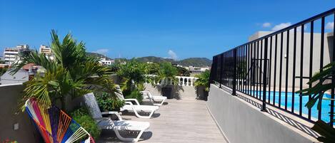 A rooftop terrace with lounge chairs, a pool, and a view of the city and hills.