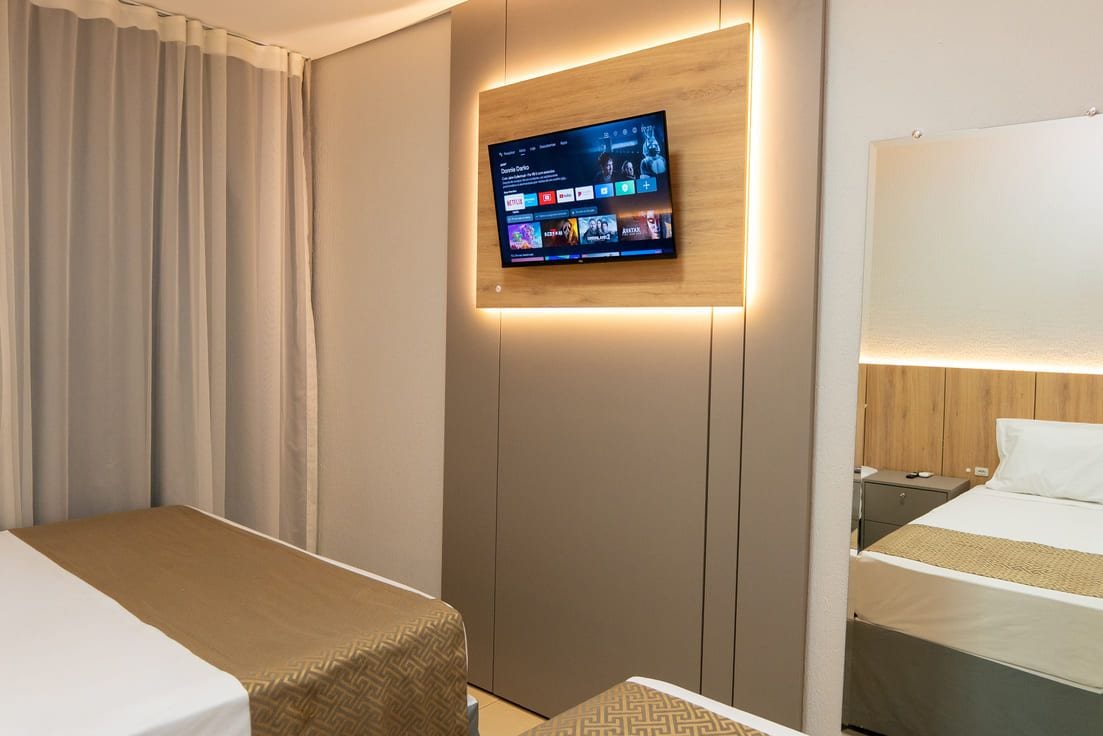 A hotel room with a flat-screen TV mounted on a wooden panel, two beds with white linens, a bedside table with a lamp, and a large window with curtains.