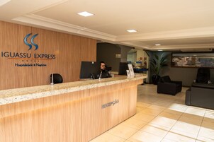 Balcão de recepção de hotel com recepcionista, parede de painéis de madeira com o nome do hotel e área de espera com assentos.