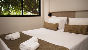A neatly made bed with white linens and patterned pillows, a window with a view of trees, and a wall-mounted light fixture. - Iguassu Express Hotel (Foz do Iguaçu)