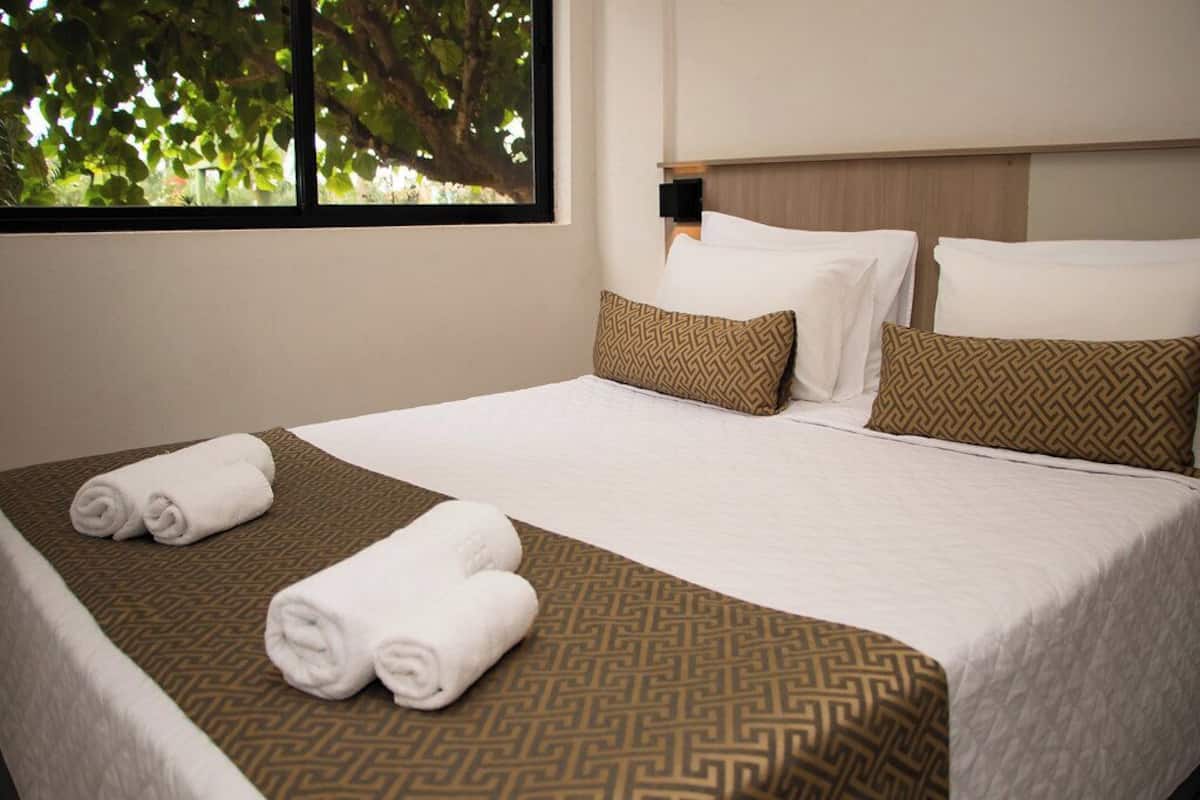 A neatly made bed with white linens and patterned pillows, a window with a view of trees, and a wall-mounted light fixture.