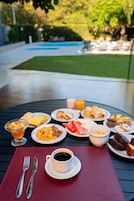 Una mesa de desayuno con diversos platos, una taza de café y una vista a una piscina y vegetación al fondo.