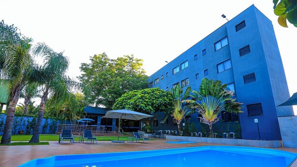 A modern building with a blue facade, a swimming pool, and outdoor seating with umbrellas. - Iguassu Express Hotel (Foz do Iguaçu)