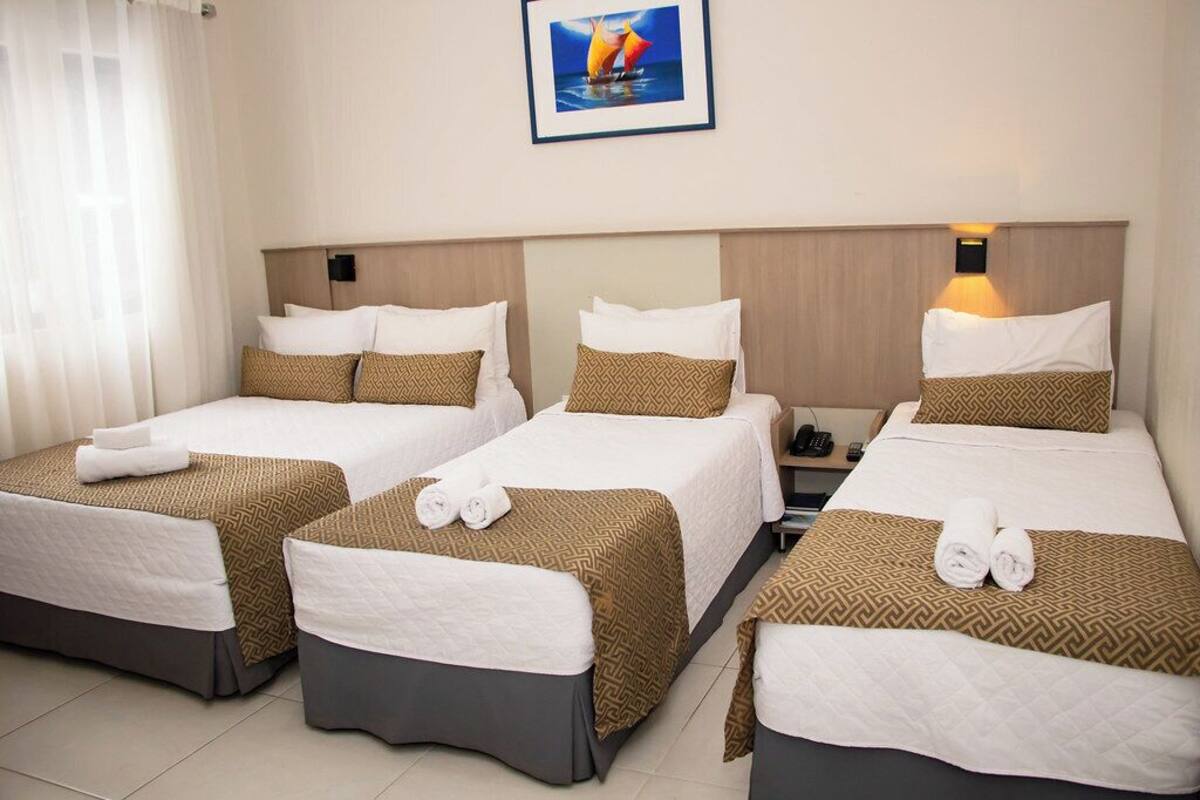 A hotel room with three single beds, each with a patterned bedspread and pillows, a bedside table with a lamp, and a framed picture on the wall.