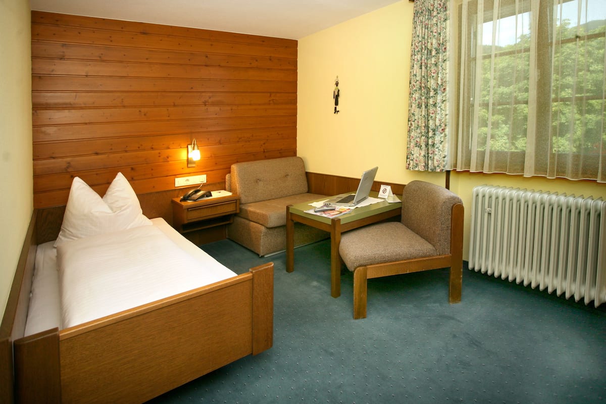 Habitación de hotel con cabecera de madera, cama con sábanas blancas, un pequeño sofá, una silla, una mesa con una computadora portátil y una ventana con vistas a árboles.