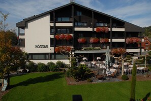 A hotel with multiple balconies adorned with red flowers, outdoor seating, and a well-maintained garden.