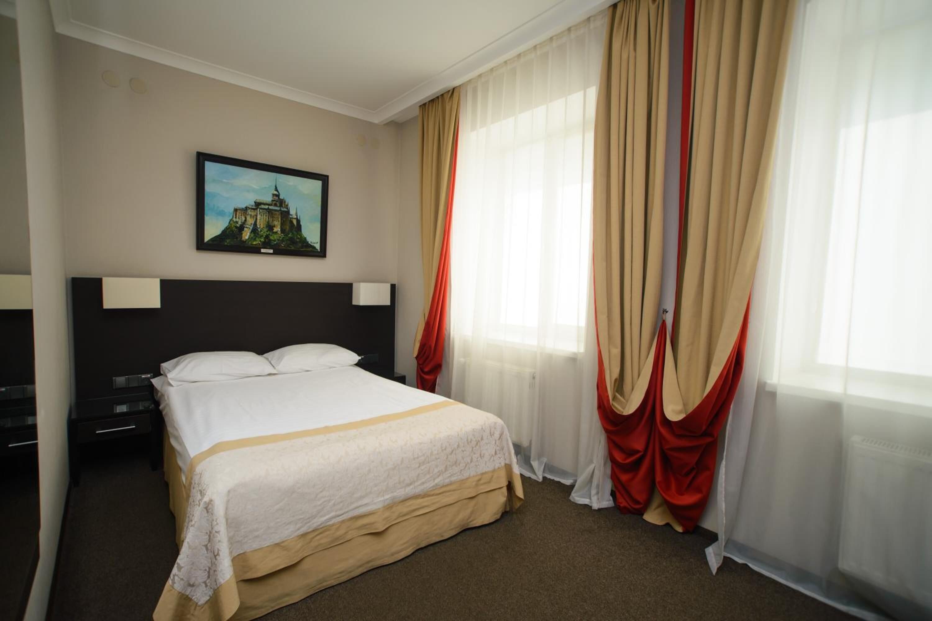 Habitación de hotel con una cama grande, una mesita de noche, una lámpara, un cuadro en la pared y cortinas traslúcidas con cortinas rojas.