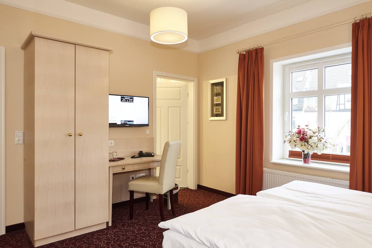 A hotel room with a bed, desk, chair, wardrobe, TV, window with curtains, and a vase with flowers.