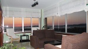 Una sala de estar con sofá, sillón y una mesita. Amplias ventanas ofrecen vistas al atardecer y a las montañas.