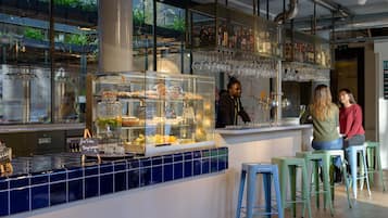 Een moderne bar met een glazen vitrine vol gebak, een wijnrek en groene barstoelen.