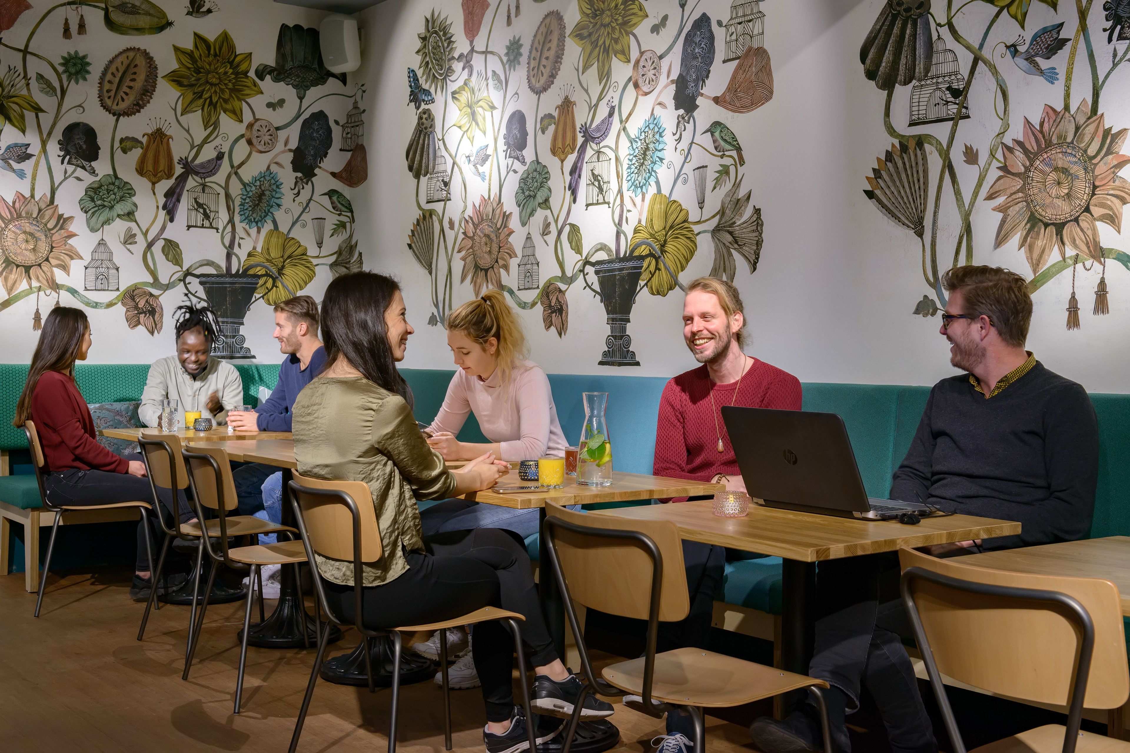 Un groupe de personnes est assis autour d’une table dans une pièce dont un mur est décoré d’une fresque représentant des illustrations botaniques.