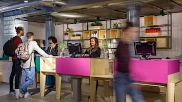 A reception desk with a pink surface, a counter, and a computer monitor. There are shelves with plants and bottles in the background.