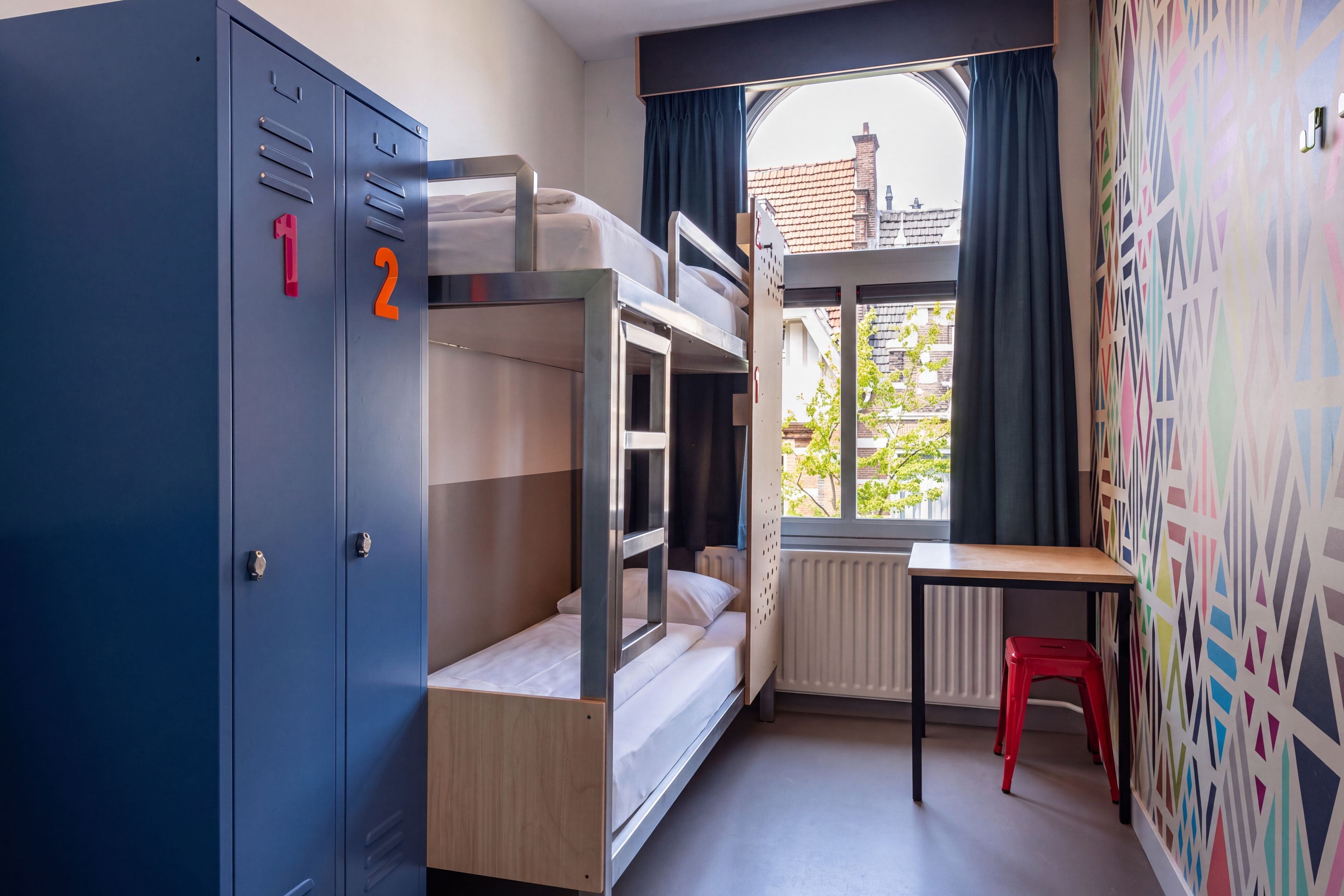 A dormitory room with bunk beds, a desk, a radiator, and a window with curtains.