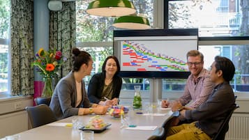 Ein Konferenzraum mit einem großen Tisch, Stühlen, einer Projektionsleinwand, die ein Gantt-Diagramm zeigt, und einem Fenster mit Vorhängen.