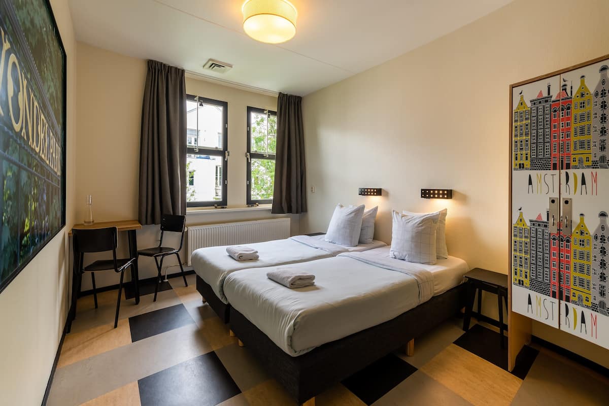 A hotel room with two beds, a desk, a chair, a window with curtains, and a cityscape artwork on the wall.