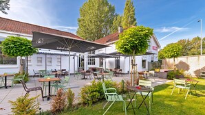 An outdoor patio area with tables and chairs, a white building with a red-tiled roof, and green trees. - Stayokay Haarlem - Hostel (Haarlem)