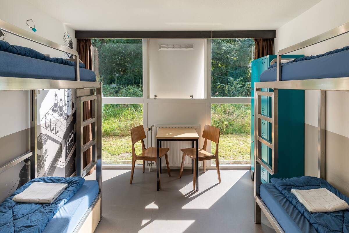 A dormitory room with bunk beds, a table, chairs, and a view of greenery outside.