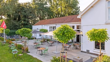 Een terras buiten met tafels en stoelen, omgeven door groen en een wit gebouw.