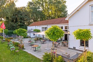 Un área de patio al aire libre con mesas y sillas, rodeada de follaje y un edificio blanco.