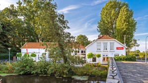 A riverside hotel with a red sign, surrounded by lush greenery and a calm water body. - Stayokay Haarlem - Hostel (Haarlem)
