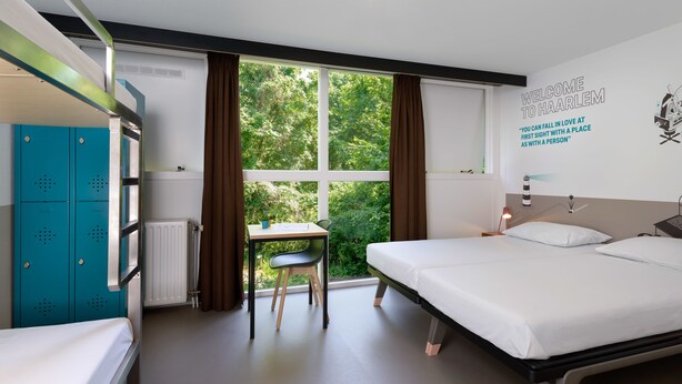 Quarto de hotel com beliches, uma escrivaninha e vista para o verde lá fora.