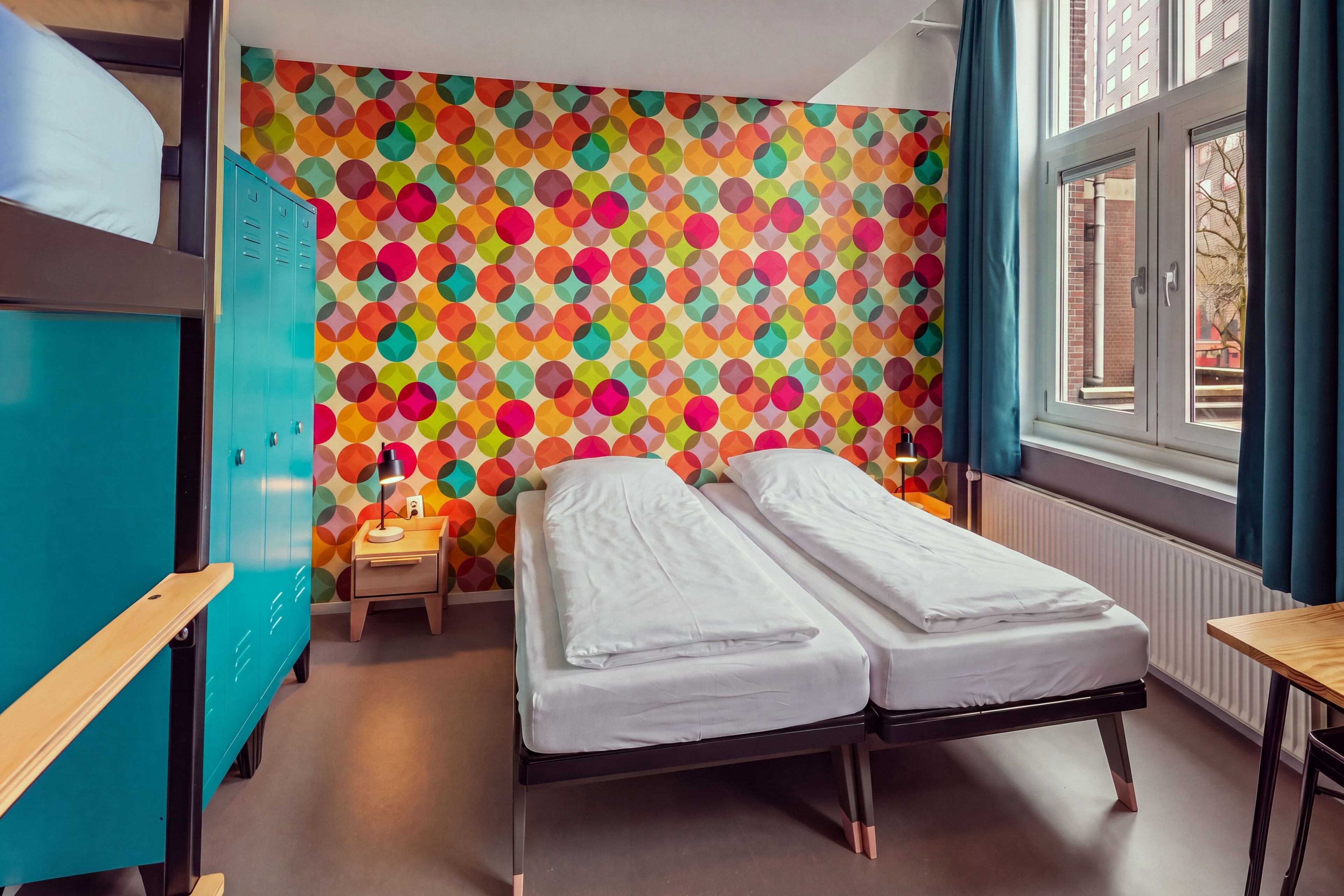 Quarto com duas camas, uma escrivaninha, um guarda-roupa e um padrão geométrico colorido na parede.