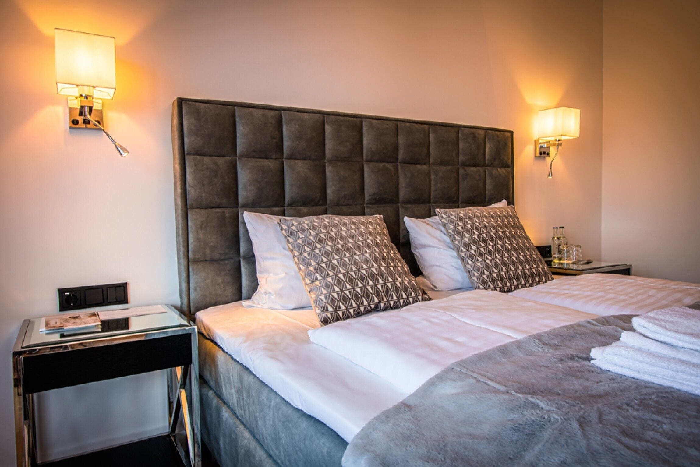 A neatly made bed with a tufted headboard, two bedside lamps, and a small table with a book and a glass.